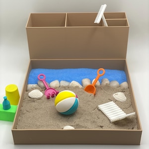Puede incluir: Una caja de madera marrón con una bandeja de arena en su interior. La bandeja contiene una pelota de playa azul y blanca, un rastrillo rosa, una pala naranja, conchas blancas y piedras blancas. Hay un rastrillo blanco y una botella de plástico verde en el compartimento superior de la caja.