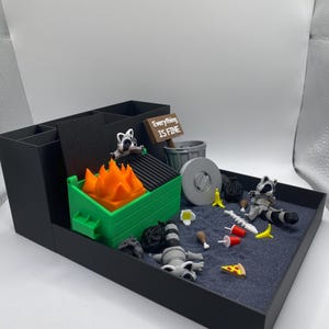 May include: A black desk organizer with a miniature diorama scene. The scene includes a green dumpster with orange flames, raccoons, trash, and a sign that reads "Everything is Fine."