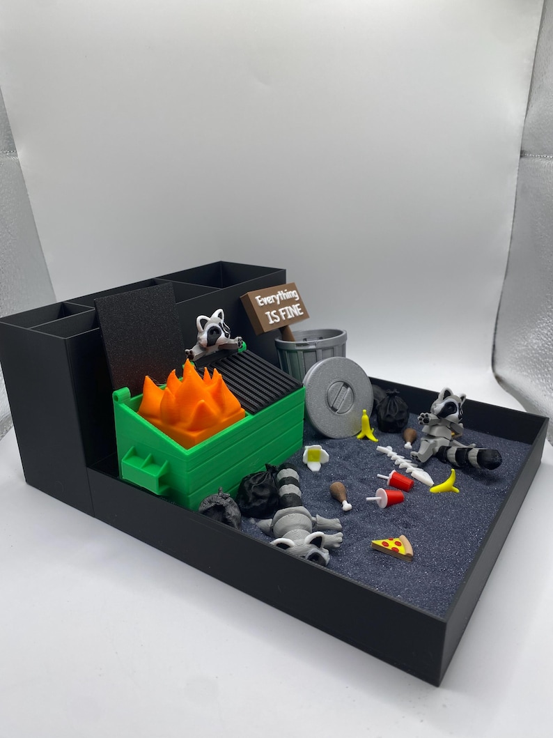 May include: A black desk organizer with a miniature diorama scene. The scene includes a green dumpster with orange flames, raccoons, trash, and a sign that reads "Everything is Fine."