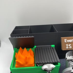 May include: A 3D-printed desk organizer featuring a green dumpster with an orange flame and a raccoon. The organizer includes a black storage compartment with multiple sections. A sign reads "Everything IS".
