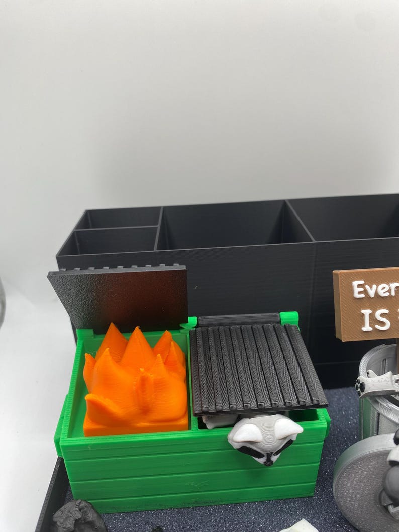 May include: A 3D-printed desk organizer featuring a green dumpster with an orange flame and a raccoon. The organizer includes a black storage compartment with multiple sections. A sign reads "Everything IS".