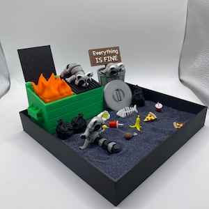 May include: A miniature diorama featuring a green dumpster with orange flames, raccoons, and trash. A sign reads "Everything is Fine." Other items include pizza slices, a banana, and a fish skeleton, all set in a black tray.