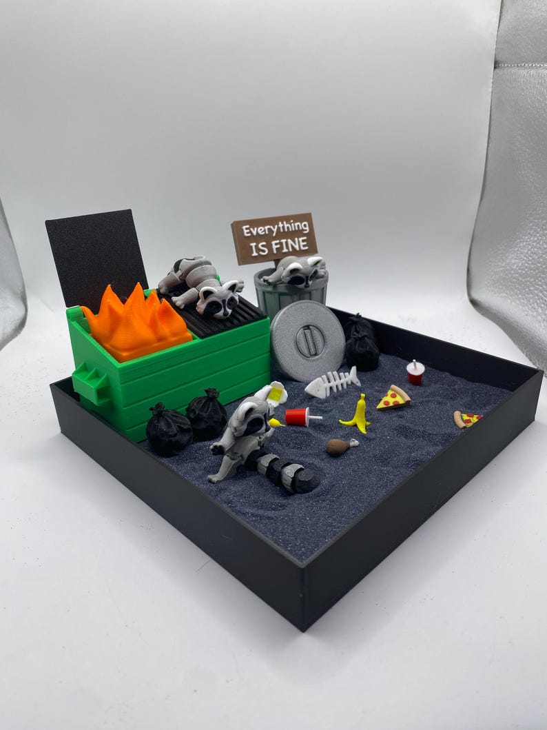 May include: A miniature diorama featuring a green dumpster with orange flames, raccoons, and trash. A sign reads "Everything is Fine." Other items include pizza slices, a banana, and a fish skeleton, all set in a black tray.
