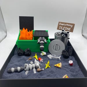 May include: A miniature diorama featuring a green dumpster with orange flames, a raccoon, and a sign that reads "Everything IS FINE." The scene includes trash bags, food items, and a gray surface.