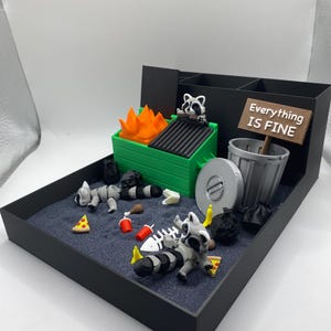 May include: A miniature diorama featuring a dumpster fire scene with raccoons, trash, and a sign that reads "Everything is Fine." The scene includes a green dumpster with orange flames, a trash can, and scattered debris.