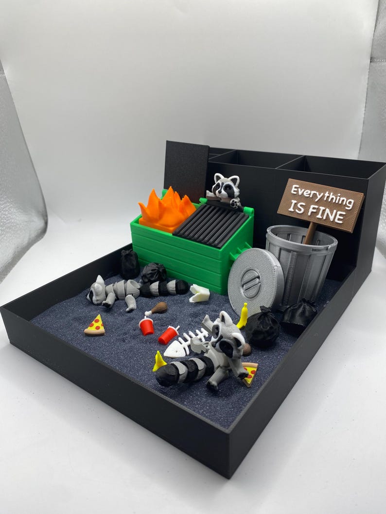 May include: A miniature diorama featuring a dumpster fire scene with raccoons, trash, and a sign that reads "Everything is Fine." The scene includes a green dumpster with orange flames, a trash can, and scattered debris.