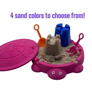 May include: A pink turtle-shaped sandbox with a lid, filled with sand and toy tools. The sandbox contains a sandcastle, an orange shovel, a blue castle mold, and a pink rake. Text above reads: "4 sand colors to choose from!"