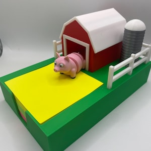 Peut inclure: Un organiseur de bureau sur le thème de la ferme avec un cochon rose sortant d'une grange rouge au toit blanc. L'organisateur comprend une zone de bloc-notes jaune, une clôture blanche et un silo gris, le tout sur une base verte.