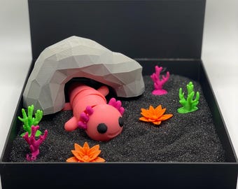 Puede incluir: Una figurita de ajolote rosa descansando debajo de una roca gris en una bandeja negra llena de arena negra. La bandeja tiene un pequeño divisor gris y un pequeño soporte negro con una varilla de metal que se extiende hacia afuera. Hay figuritas de coral y flores verdes, rosas y naranjas esparcidas alrededor del ajolote.