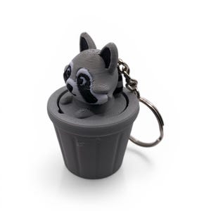 Puede incluir: Llavero gris con un mapache impreso en 3D asomando de un cubo de basura a juego. El mapache tiene marcas en blanco y negro. El llavero incluye un anillo plateado para sujetar a llaves o bolsos.