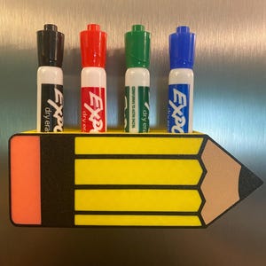 May include: A yellow pencil-shaped magnetic holder with a pink eraser end and a dark gray tip, holding four Expo dry erase markers in black, red, green, and blue. The holder features black vertical stripes and is attached to a metallic surface.