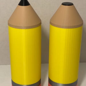 Puede incluir: Dos recipientes cilíndricos con forma de lápices amarillos, con puntas cónicas de color beige, extremos negros y una banda gris que separa el cuerpo amarillo de la base rosa. Un lápiz tiene punta afilada y el otro una parte superior plana.