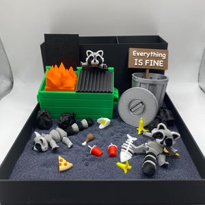 May include: A miniature diorama featuring a raccoon-themed scene. A green dumpster with orange flames, a trash can, and a sign that reads "Everything IS FINE" are present. Raccoons are depicted with various items, including a fish skeleton and pizza.