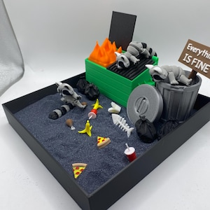 May include: A miniature diorama featuring a green dumpster with orange flames and sleeping raccoons. The scene includes trash, pizza slices, and a sign that reads "Everything is Fine."