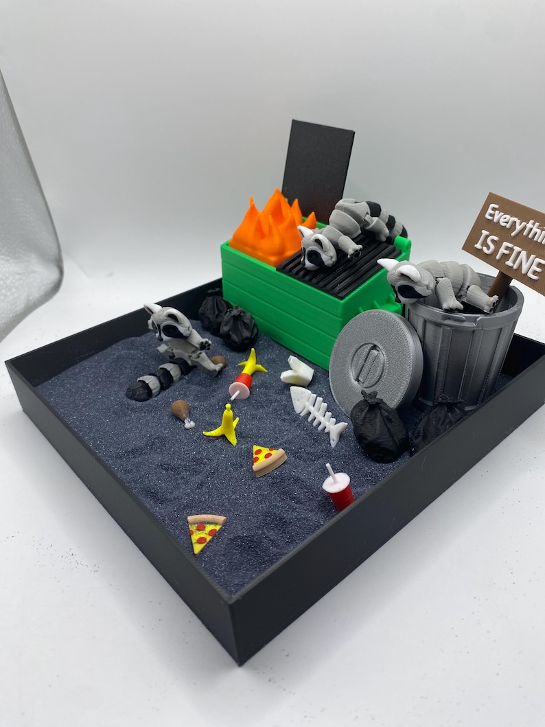 May include: A miniature diorama featuring a green dumpster with orange flames and sleeping raccoons. The scene includes trash, pizza slices, and a sign that reads "Everything is Fine."