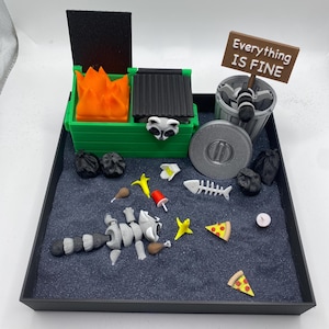 May include: A miniature diorama featuring a green dumpster with orange flames, a trash can, and a sign that reads "Everything IS FINE." A raccoon, pizza slices, and other trash are scattered on a dark surface within a black tray.