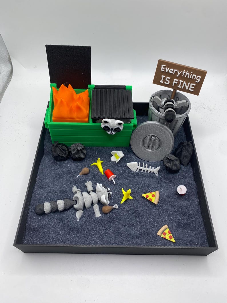 May include: A miniature diorama featuring a green dumpster with orange flames, a trash can, and a sign that reads "Everything IS FINE." A raccoon, pizza slices, and other trash are scattered on a dark surface within a black tray.
