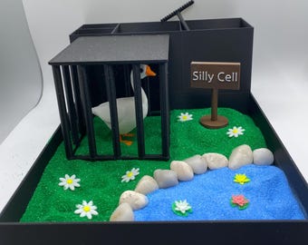 Puede incluir: Un diorama caprichoso con un ganso blanco en una jaula negra con la etiqueta "Silly Cell". La escena incluye un estanque azul, una zona de césped verde, margaritas blancas y piedras. Una caja negra con compartimentos está en el fondo.