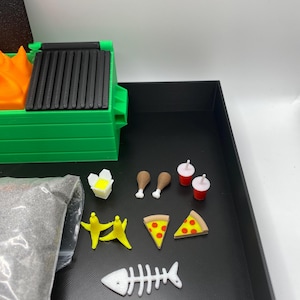 May include: A miniature green dumpster with a black lid and orange flames, next to a bag of gray material. Also includes miniature food items: pizza slices, chicken legs, bananas, a fish skeleton, and drinks.