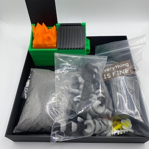 May include: A miniature diorama set featuring a green dumpster with orange flames, a black lid, and a gray grill. The set includes a bag of gray sand, a bundle of black and white objects, and a small sign that reads "everything IS FINE".