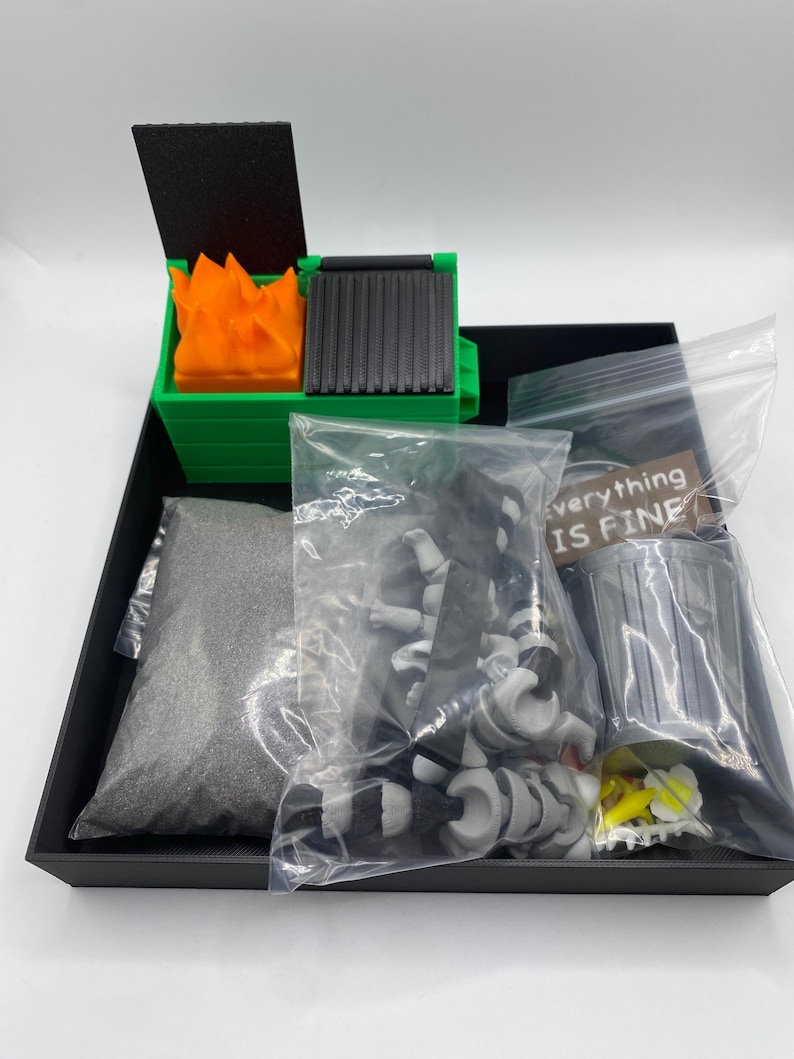 May include: A miniature diorama set featuring a green dumpster with orange flames, a black lid, and a gray grill. The set includes a bag of gray sand, a bundle of black and white objects, and a small sign that reads "everything IS FINE".