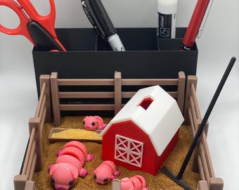 Puede incluir: Un organizador de escritorio negro con tijeras rojas, un marcador negro, una barra de pegamento y un bolígrafo rojo. Una escena de granja en miniatura presenta un granero rojo y blanco, una valla de madera y figuras de cerdos rosas.