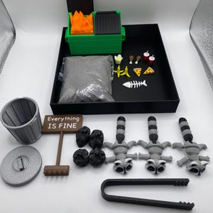 May include: A miniature diorama set featuring a green dumpster with flames, a trash can, a sign that reads "Everything IS FINE", raccoons, and food items. The set includes a black tray with a bag of gray material.