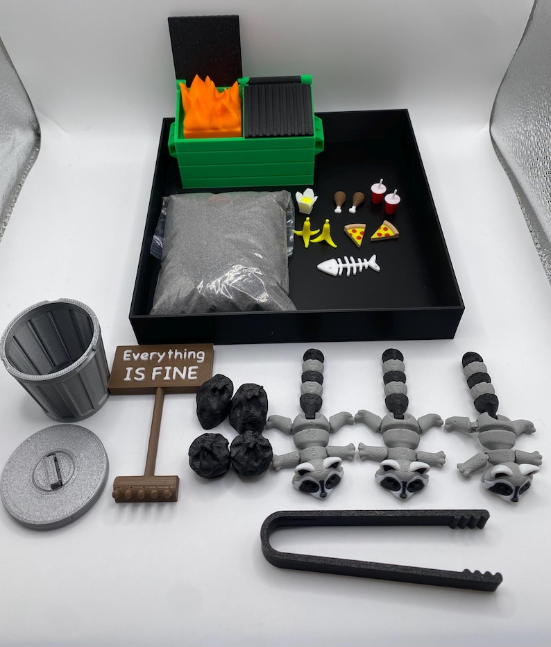 May include: A miniature diorama set featuring a green dumpster with flames, a trash can, a sign that reads "Everything IS FINE", raccoons, and food items. The set includes a black tray with a bag of gray material.