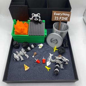 May include: A whimsical diorama featuring a black organizer with compartments, a green dumpster with orange flames, and a sign that reads "Everything IS FINE." Includes miniature trash, a raccoon, and food items.