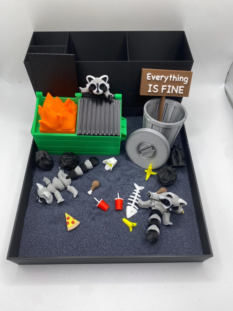 May include: A whimsical diorama featuring a black organizer with compartments, a green dumpster with orange flames, and a sign that reads "Everything IS FINE." Includes miniature trash, a raccoon, and food items.