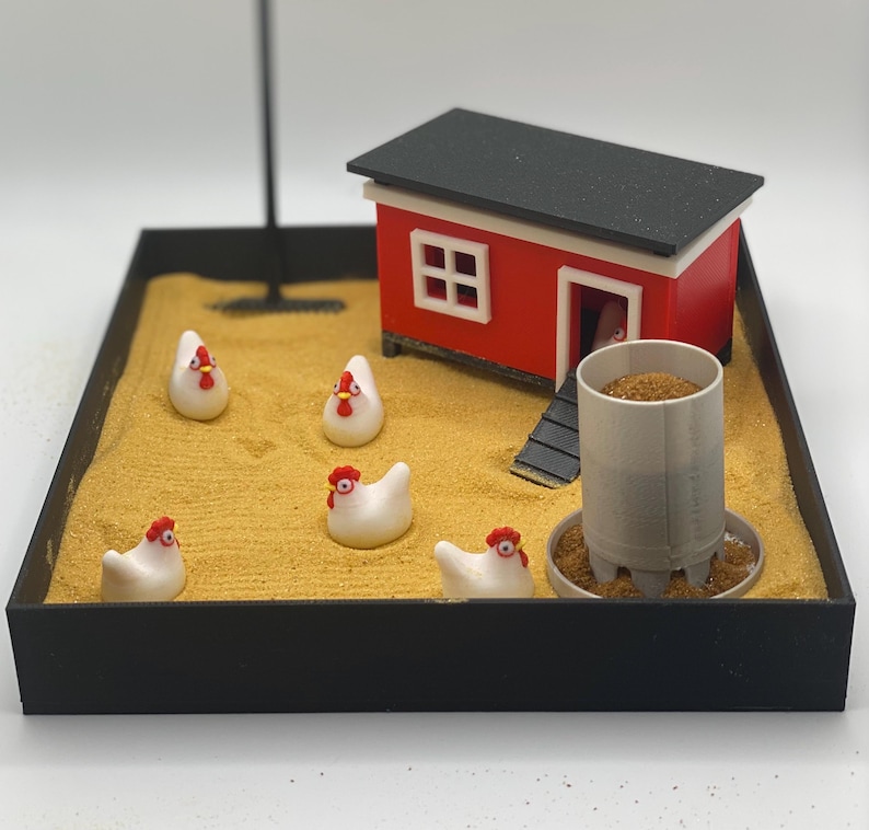 Może przedstawiać: Miniaturowy czerwony kurnik z czarnym dachem stoi na piaskowej tacy. Pięć białych ceramicznych kur z czerwonymi grzebieniami rozrzuconych wok&oacute;ł kurnika. Szary metalowy karmnik z brązowym pokarmem znajduje się przed kurnikiem.