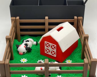 Puede incluir: Un organizador negro con un granero rojo, dos vacas blancas y negras y un área verde cubierta de hierba encerrada por una valla marrón. El organizador tiene un rastrillo en el lateral.