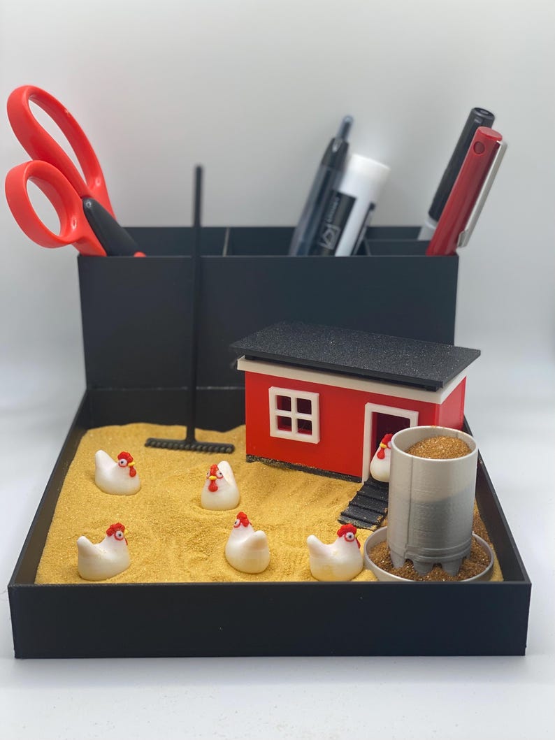 Może przedstawiać: Czarny organizer biurkowy z miniaturowym kurnikiem w kolorze czerwonym i białym, czarnym grabiami i pięcioma białymi ceramicznymi kurami z czerwonymi grzebieniami. Organizer ma przegrody na długopisy, nożyczki i inne artykuły biurowe.