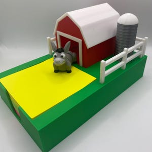 May include: A farm-themed desk tidy with a red barn and white roof, a grey silo, and a white fence. A smiling donkey figurine stands on a yellow field. The base is green.