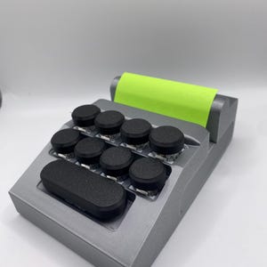 Puede incluir: Un dispositivo rectangular gris con ocho botones redondos negros y un botón rectangular negro. Una nota adhesiva verde neón está adherida a la parte superior. El dispositivo está sobre un fondo blanco.