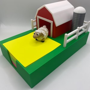 Puede incluir: Un organizador de escritorio con temática de granja con un granero rojo y techo blanco, un silo y una valla blanca. Un bloc de notas amarillo se encuentra sobre una base verde, con una pequeña figura de oveja. Diseñado para notas adhesivas.