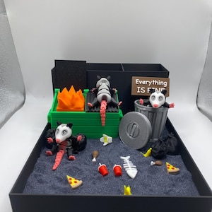 May include: A miniature diorama featuring opossums in a trash-themed setting. One opossum is grilling, another is in a trash can, and a third is lounging. The scene includes food items and a sign that reads "Everything IS FINE."