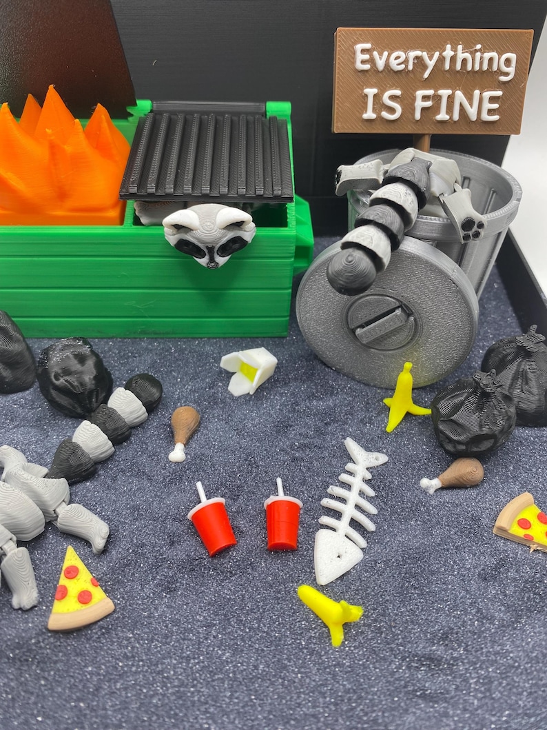 May include: A miniature diorama featuring a green dumpster with a raccoon head, a trash can with a raccoon, and various food items. A sign reads "Everything IS FINE". Includes pizza, fish bones, and soda cups.
