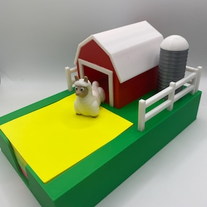 May include: A miniature farm scene with a red barn, white roof, and a silo. A yellow field and a white fence are also present. A small, white llama figure stands near the barn. The base is green.