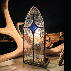 Könnte beinhalten: Ein Buntglasfenster in Bogenform mit einem blauen Diamanten in der Mitte. Das Glas hat ein strukturiertes, wellenförmiges Aussehen. Das Fenster ist vor einem dunklen Hintergrund mit einem Hirschgeweih und dekorativen Elementen platziert.