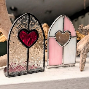 Puede incluir: Dos adornos de ventana de vidrieras. Uno es dorado y rojo con forma de corazón, el otro es rosa y blanco con forma de corazón. Ambos tienen un borde negro y forma arqueada.