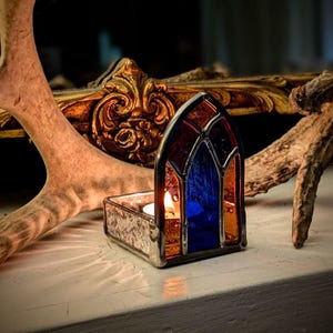 Könnte beinhalten: Ein Kerzenhalter aus Buntglas mit gotischem Bogen-Design. Das Glas ist eine Mischung aus Blau, Orange und Rot. Eine brennende Kerze befindet sich im Halter und wirft einen warmen Schein. Der Halter steht auf einer weißen Oberfläche, mit Geweih und einem dekorativen Goldrahmen im Hintergrund.
