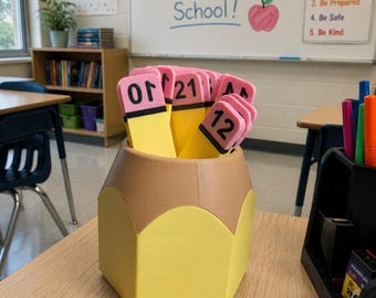 May include: A yellow and tan pencil-shaped desk organizer holding several flat, pencil-themed markers. Each marker features a pink eraser top, a black band, and a yellow body with black numbers or letters printed on the front, including 21 and 12.