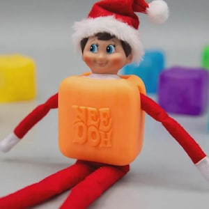 May include: A small elf doll wearing a red and white Santa hat, featuring a smiling face with blue eyes. The doll is wearing a bright orange square-shaped Nee Doh sensory toy around its torso, with long red fabric arms and legs ending in white hands.