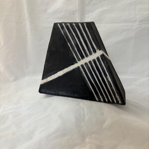 May include: A black, geometric vase with white linear accents. The vase has a triangular shape with a flat base and a wide opening. White lines run diagonally across the front, creating a striking contrast against the black surface.