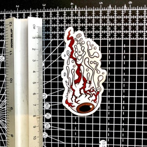 The image shows a melting eyeball sticker on a cutting mat alongside a ruler to illustrate its size.