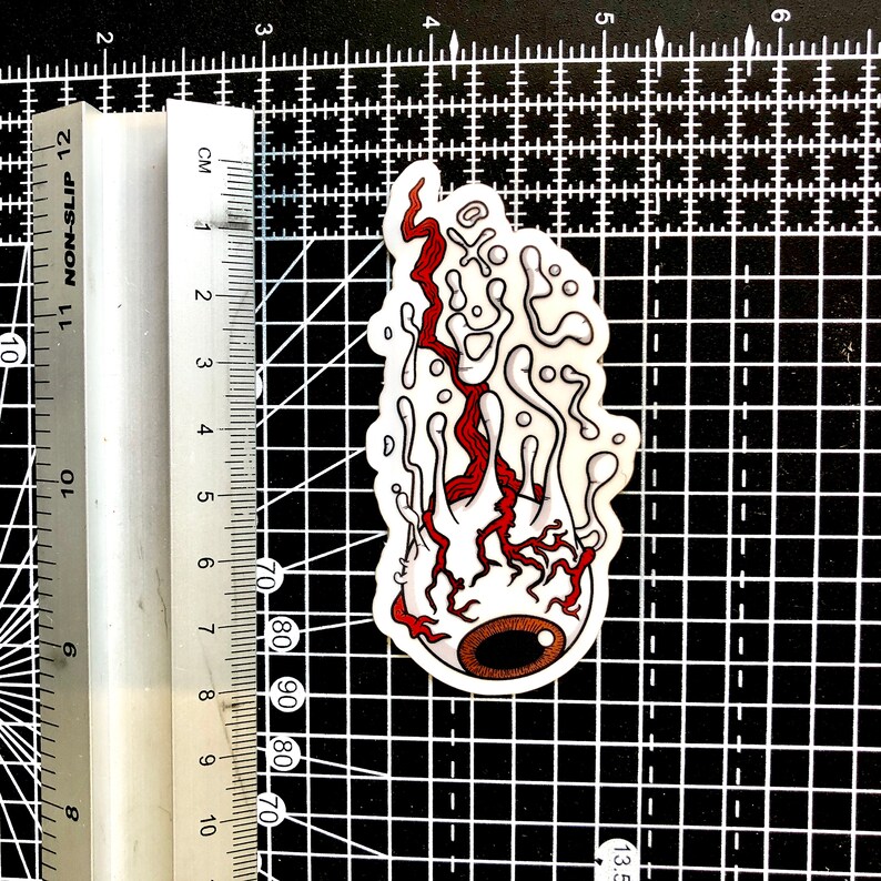 The image shows a melting eyeball sticker on a cutting mat alongside a ruler to illustrate its size.