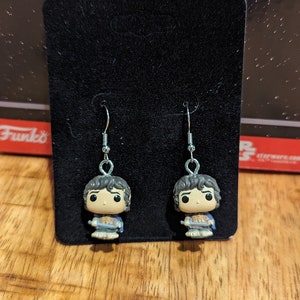 May include: A pair of silver dangle earrings featuring a small, detailed figurine of a character from the Lord of the Rings. The figurine is wearing a blue tunic and brown pants. The earrings are displayed on a black background.