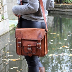 May include: A brown leather satchel bag with a flap closure and multiple buckles. The bag is worn over the shoulder of a person wearing a grey sweater and black trousers.