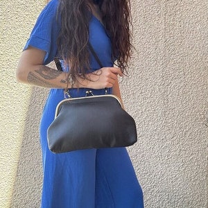 Peut inclure: Une pochette en satin noir avec une fermeture à fermoir dorée et une bandoulière en chaîne. Le sac est tenu par une personne portant une combinaison bleue.